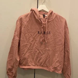 Rosa hoodie från Divided - Säljer en supermysig rosa hoodie från Divided. Den har texten 'B.A.B.E.S' tryckt på framsidan. Perfekt för chilliga dagar eller när du bara vill vara bekväm. Den är i bra skick och redo för en ny ägare!