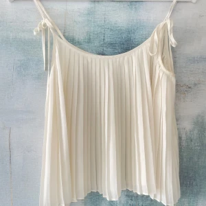 Vit plisserad topp med knytband - Säljer en superfin vit plisserad topp med knytband på axlarna. Den är perfekt för sommardagar och har en luftig och elegant stil. 