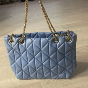 Blå quiltad axelväska med guldkedja - Säljer en superfin blå axelväska med quiltat mönster. Väskan har en elegant guldkedja som axelrem och är perfekt för både vardag och fest. Den är rymlig och stilren, vilket gör den till ett måste i garderoben!