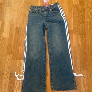 Blå jeans med vita ränder och knytband - Säljer ett par coola lågmidjade blå jeans från Edikted i storlek XS. De har vita ränder längs sidorna och snygga knytband vid anklarna. Helt oanvända och köpta för 1600