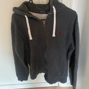 Grå hoodie från Polo Ralph Lauren - Säljer en snygg grå hoodie från Polo Ralph Lauren. Den har en dragkedja framtill och en klassisk huva med vita snören. Perfekt för en avslappnad stil och passar bra till både höst och vår. Den är i bra skick. Tveka inte att höra av er vid funderingar