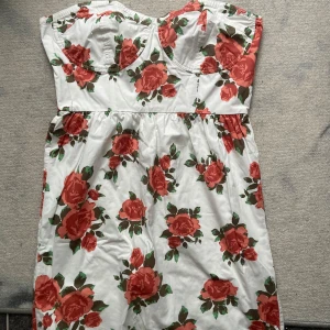 Blommig axelbandslös klänning - Säljer en superfin axelbandslös klänning med ett härligt blommigt mönster. Klänningen är vit med stora röda rosor och gröna blad. Perfekt för sommarens alla fester! 🌹
