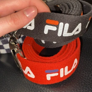 Fila-skärp - Fila-skärp. Ett rött och ett i svart. Båda är köpta i Japan och är ca 45 cm långa. På det röda skärpet är den blå färgen i loggan mörkare i verkligheten än på bild.140 kr/st
