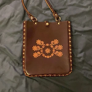 Gammaldags brun/lila handväska  - Jättecool väska köpt på second hand! Formen är som en tygpåse, den är i något slags läder! Färgen är brun/lila och blom-”trycket” är ljusbrunt. Nitar längs alla kanter och en knapp att stänga med, datorn får plats perfekt!❤️‍🔥❤️‍🔥❤️‍🔥budgiving vid stort intresse!