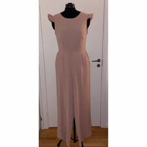 Rosa Jumpsuit med Knytrygg - Rosa jumpsuit med knytdetalj i ryggen och liten volang vid axeln. Aldrig använd, nypris 600kr