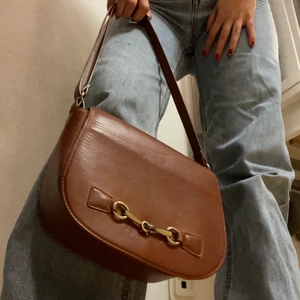 Brun hanväska i vintagelook - En söt och sjukt praktisk handväska i fakeläder med fint metallspänne. Bandet går att förlänga och väskan rymmer mycket; mobil, powerbank, hörlurar och tillockmed en pocketbok. Klassisk look. Den är väl använd men håller nog ett tag till. DM för fler bilder/ frågor. Öppen för att diskutera priset. 