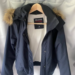 Marinblå jacka från Hollister i storlek M - Marinblå jacka från Hollister i storlek M i mycket fint skick. Luva med fuskpäls. Pris 100kr