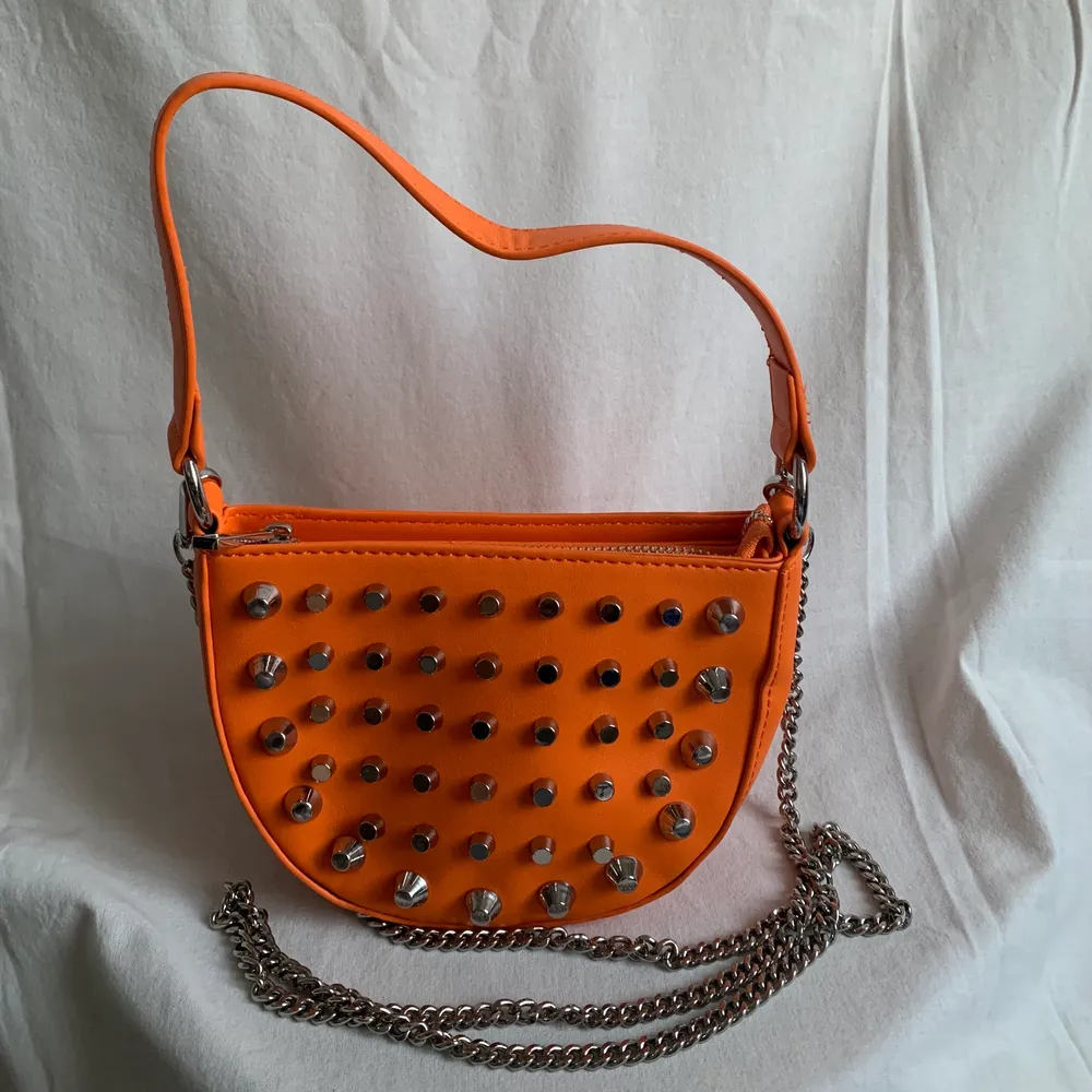 Orange liten väska från asos med silvriga nitar på ena sidan, axelremmen är en silvrig kedja. Både kedjan och den korta remmen går att ta av. Helt oanvänd! . Väskor.