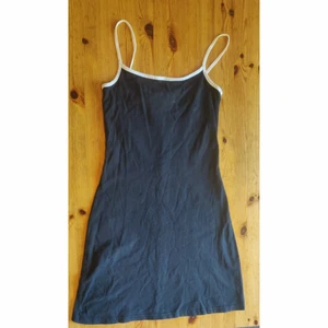 Svart klänning med vit linning - En svart stretchig klänning med vita kontrastsömmor vid halslinningen och vid axelband. Tajt upptill och sedan lätt utsvängd nertill, snygg som den är eller till exempel över en tjocktröja. Tyget har lite tecken på användning men inga fläckar eller dylikt. Storlek S.  Avhämtning i Stockholm eller skickas mot frakt. 