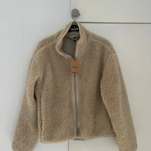 Beige teddyjacka från Weekday - Mysig beige teddyjacka från Weekday med dragkedja framtill. Perfekt för kyliga dagar med sin fluffiga och varma design. Jackan har en enkel och stilren look som passar till många olika outfits.