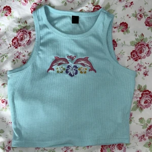 Turkos ribbad linnetopp med delfinbroderi - Säljer en söt turkos ribbad linnetopp med ett färgglatt broderi av delfiner och blommor på framsidan. Relativt kort, och lappen går nästan inte att läsa. Perfekt för sommardagar! 💙