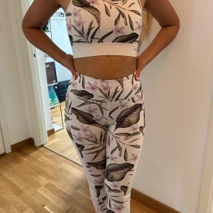 Blommig träningsset i vitt och rosa - Snyggt träningsset med en croppad topp och matchande leggings. Setet har ett blommigt mönster i vitt, rosa och brunt. Toppen har en öppen rygg och är ärmlös, perfekt för träning.