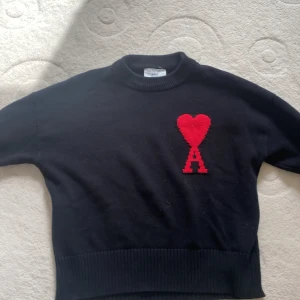 Svart tröja från Ami Alexandre Mattiussi - Säljer en svart tröja från Ami Alexandre Mattiussi med ett rött hjärta och bokstaven A på bröstet. Tröjan har långa ärmar och en ribbad nederkant. Perfekt för en stilren look.