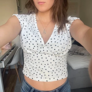 Vit prickig croppad topp - Säljer en söt vit topp med svarta prickar och v-ringning. Toppen är croppad och har korta ärmar, perfekt för sommardagar. Den är både stilren och lekfull, vilket gör den lätt att matcha med olika outfits.