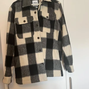 Rutig overshirt från Zara - Snygg rutig overshirt från Zara i svart och vit. Jackan har långa ärmar och knappar framtill samt två bröstfickor med lock. I fint skicka, sömmar och knappar är hela och inga nopprar eller dylikt slitage