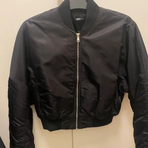 Svart bomberjacka från Gina Tricot - Snygg svart bomberjacka från Gina Tricot med dragkedja framtill. Jackan har en klassisk bomberstil med ribbade muddar vid ärmslut och nederkant. Perfekt för en cool och avslappnad look. Ordinarie pris är ca 250kr. Fint skick utan slitage. 