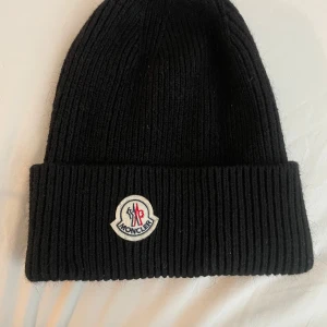 Moncler mössa - Snygg svart ribbad mössa från Moncler med broderad logotyp framtill. Perfekt för att hålla värmen under kyliga dagar. Stilren design som passar till det mesta.