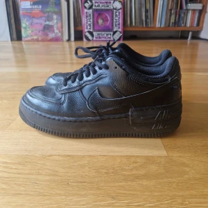 Svarta Nike Air Force 1 Shadow sneakers - Säljer ett par klassiska svarta Nike Air Force sneakers i läder. Skorna har en stilren design med snörning och Nike-logga på sidan. Perfekta för en casual look och bekväma att bära hela dagen.