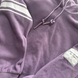 Lila långklänning med huva - Mysig lila långklänning med huva och snörning. Klänningen har långa ärmar med vita ränder och en patchdetalj på ärmen. Perfekt för en avslappnad stil.