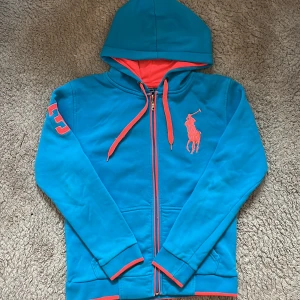 Blå hoodie från Ralph Lauren - Säljer en snygg blå hoodie från Ralph Lauren med orange detaljer. Tröjan har en dragkedja framtill och en stor broderad logga på bröstet. Den har långa ärmar och en bekväm passform. Perfekt för en avslappnad stil!