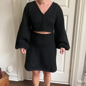 Svart klänning från Adoore - Snygg stickadsvart klänning från Adoore med v-ringning och knappar framtill. Klänningen har långa ärmar och en croppad toppdel som ger en modern look. Helt ny aldrig använd