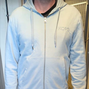 Ljusblå hoodie från Michael Kors - Säljer en stilren ljusblå hoodie från Michael Kors med dragkedja och huva. Perfekt för en avslappnad stil. KORS-logga på bröstet ger en exklusiv touch. Passar bra till både jeans och joggers. I väldigt gott skick, använts några enstaka gånger. Storlek: Medium. Pris: 600kr