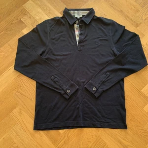 Svart pikétröja från Burberry - Priset kan diskuteras. Snygg svart pikétröja från Burberry med klassisk krage och knappar. Tröjan har ett diskret broderat märke på bröstet och ikoniskt rutigt mönster på insidan av kragen. Perfekt för en stilren look. 