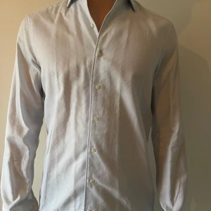 Ljusblå långärmad skjorta - Snygg ljusblå långärmad skjorta med diskret mönster och klassisk krage. Perfekt för en stilren look. Skjortan har knappar framtill och är tillverkad i ett lätt och bekvämt material.