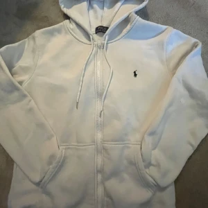 Vit hoodie från Polo Ralph Lauren - Säljer en stilren vit hoodie från Polo Ralph Lauren med dragkedja och den klassiska loggan broderad på bröstet. Den har en justerbar huva med snören och praktiska fickor framtill.