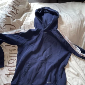 Blå hoodie från GANT - Säljer en snygg blå hoodie från GANT med vita och röda ränder längs ärmarna. Tröjan har en klassisk känguruficka och en bekväm huva. Perfekt för en avslappnad stil.