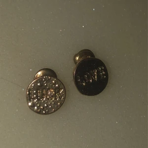 Guldörhängen med text - Säljer ett par runda guldörhängen med ingraverad text. Ena örhänget har ordet 'LUCK' och det andra 'LOVE'. Perfekta för att ge en subtil men meningsfull touch till din stil.