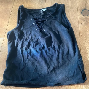Svart ärmlös topp från H&M Divided - Säljer en svart ärmlös topp från H&M Divided. Toppen har en snygg snörning framtill som ger en cool detalj. Perfekt för en avslappnad stil eller att klä upp med accessoarer. Passar bra till jeans eller kjol.