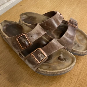 Bruna sandaler från Birkenstock - Säljer ett par klassiska bruna sandaler från Birkenstock med dubbla justerbara remmar och metallspännen. Perfekta för sommarens alla äventyr. Sandalerna har en bekväm fotbädd och slitstark sula.