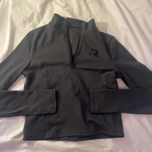 Svart långärmad tröja med dragkedja - Säljer en svart långärmad tröja med dragkedja och hög krage. Tröjan är tillverkad av 90% nylon och 10% spandex, vilket ger en stretchig och bekväm passform. Perfekt för träning eller vardagsbruk.