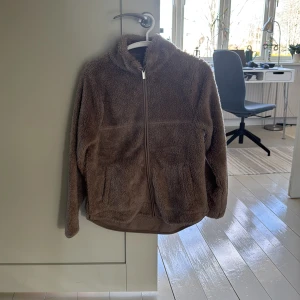 Brun teddyjacka från 157 - Mysig brun teddyjacka från 157 med dragkedja framtill. Perfekt för kyliga dagar med sin fluffiga och varma design. Jackan har långa ärmar och en hög krage för extra värme.