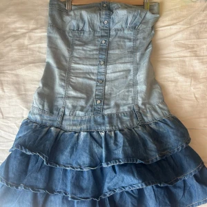 Jeansklänning med volanger - Snygg axelbandslös jeansklänning med volanger i olika blå nyanser. Klänningen har knappar framtill och en smockdetalj på baksidan för bättre passform. Perfekt för en avslappnad stil med en touch av denim.