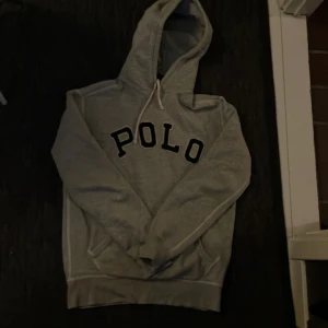 Grå hoodie från Polo Ralph Lauren - Säljer en grå hoodie från Polo Ralph Lauren med stor svart text på framsidan. Hoodien har en klassisk design med i huvan och ribbade muddar vid ärmslut och nederkant. Perfekt för en avslappnad stil. Passar S och M