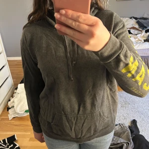 Grå hoodie  - Säljer en grå hoodie från American Eagle med gul text längs ärmen. Väl använd och nopprig