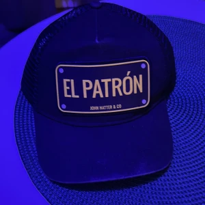Svart keps med texten 'El Patrón' - Snygg svart keps från John Hatter & Co med texten 'El Patrón' på framsidan. Kepsen har ett nätmaterial på baksidan för bättre ventilation och en klassisk böjd skärm. Perfekt för en cool och avslappnad stil.