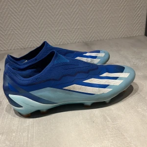 Blå fotbollsskor från Adidas X/ Addidas högsta model. - Snygga blå fotbollsskor från Adidas med vita detaljer och en strömlinjeformad design. Skorna har en rund tå och är perfekta för fotbollsplanen. De är tillverkade i syntetmaterial och har en tight passform för optimal kontroll.