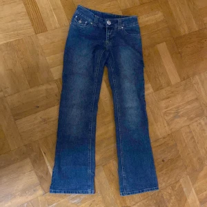 Blå bootcut jeans med dekorativa detaljer - Snygga blå bootcut jeans med dekorativa stenar på fickorna och knappen. Perfekta för en avslappnad stil med en touch av glamour. De har en klassisk femficksdesign och är tillverkade i ett bekvämt denimtyg. Jeansknappen är dock trasig men går att fixas själv.