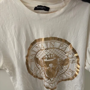 Vit t-shirt från Balmain med guldtryck - Säljer en stilren vit t-shirt från Balmain med ett iögonfallande guldtryck på framsidan. T-shirten har en klassisk rund hals och korta ärmar. Perfekt för dig som vill ha en touch av lyx i vardagen.