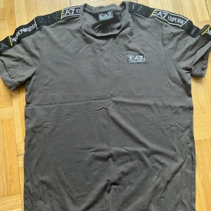 Svart t-shirt från Emporio Armani - Snygg svart t-shirt från Emporio Armani med EA7-logga på bröstet och längs ärmarna. Perfekt för en stilren look med en touch av lyx. Kortärmad och tillverkad i mjukt material för extra komfort. Storlek S