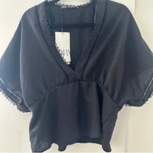 Svart blus från Zara - Snygg svart blus från Zara med v-ringning och korta ärmar. Blusen har fina spetsdetaljer längs kanterna och en lös passform som ger en avslappnad look.