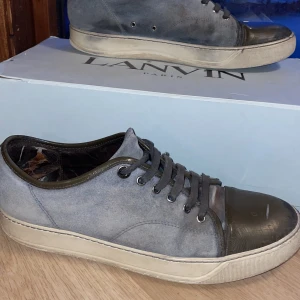 Grå/gröna sneakers från Lanvin - Snygga grå sneakers från Lanvin med en unik design. Skorna har en rund tå och snörning framtill. De är tillverkade i mocka med detaljer i skinn, vilket ger en stilren look. Perfekta för dig som vill ha något exklusivt och bekvämt.