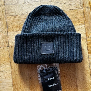 Grå stickad mössa från Acne Studios - Snygg svart stickad mössa från Acne Studios med ett stilrent broderat ansikte på framsidan. Perfekt för att hålla värmen under kyliga dagar. Mössan har en ribbad design som ger en skön passform. Färg: Charcoal