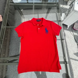 Röd pikétröja från Ralph Lauren - Säljer en snygg röd pikétröja från Ralph Lauren med en stor blå broderad logga på bröstet. Tröjan har en klassisk krage och knappar framtill. Perfekt för en stilren look!