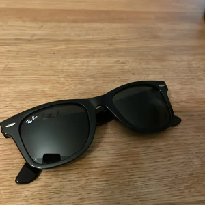 Svarta solglasögon från Ray-Ban - Snygga svarta Wayfarer-solglasögon från Ray-Ban. Klassisk design med robust båge och mörka linser. Perfekta för en stilren look året runt. Fodral medföljer ej.