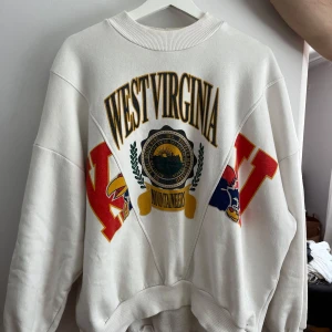 Vit sweatshirt med West Virginia-tryck från chiquelle - Säljer en vit sweatshirt med tryck av 'West Virginia' och färgglada detaljer i rött och gult. Tröjan har långa ärmar och en rund halsringning. Perfekt för en avslappnad stil.
