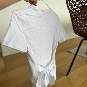 Vit t-shirt från Zara med sidenrosett på ryggen - Säljer en stilren vit topp med en sidenband baktill. Den är aldrig använd, därav vecken/skrynklorna på bilderna. Säljer för 130 kronor. 